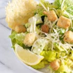 Bowl of caesar salad with grated parmesan cheese, croutons, a slice of lemon, and a parmesan cheese crisp
