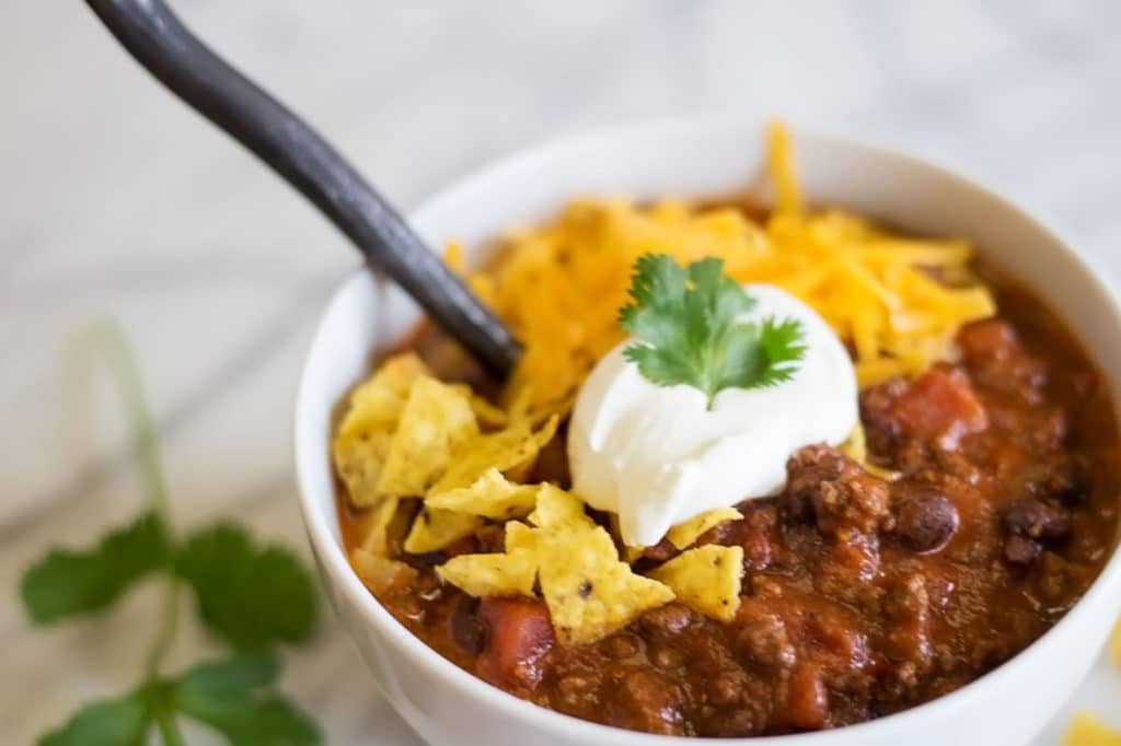 White bowl of chili garnished with sour cream, crunched up tortilla chips, and grated cheddar cheese