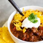 A white bowl of chili garnished with sour cream, crushed tortilla chips, and grated cheddar cheese