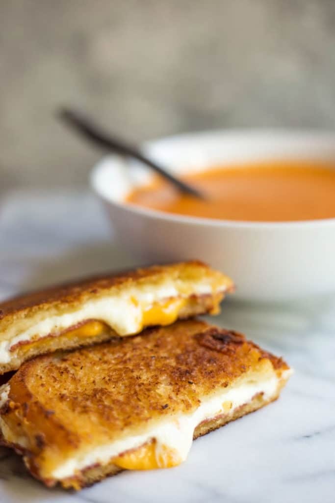 A grilled cheese and bacon sandwich cut in half in front of a bowl of tomato soup
