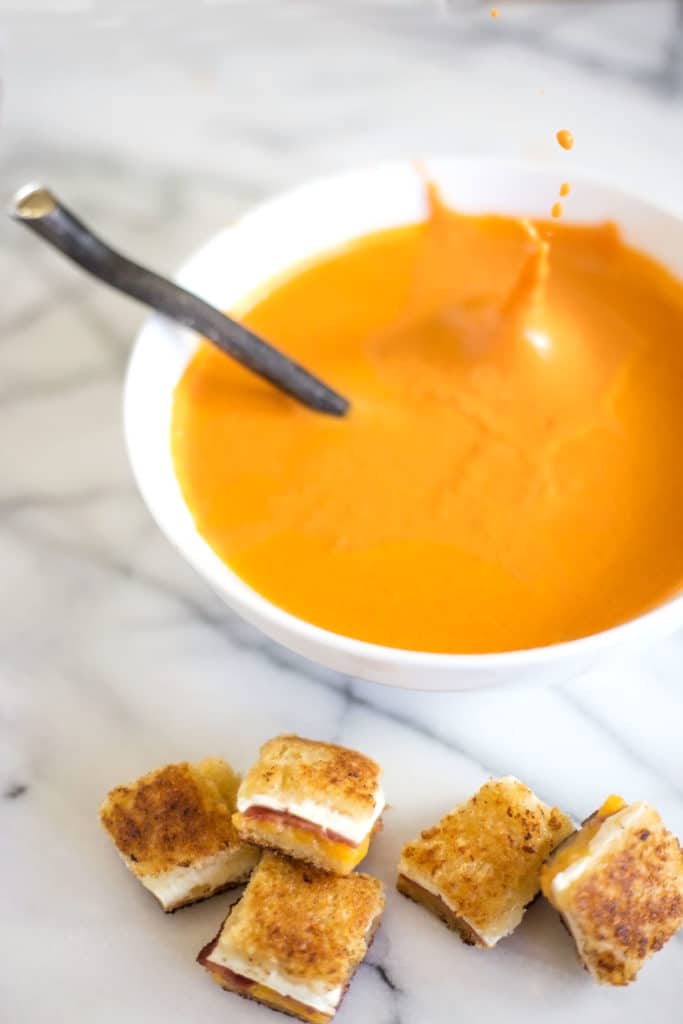 Bowl of tomato soup with a splash surrounded by grilled cheese and bacon croutons
