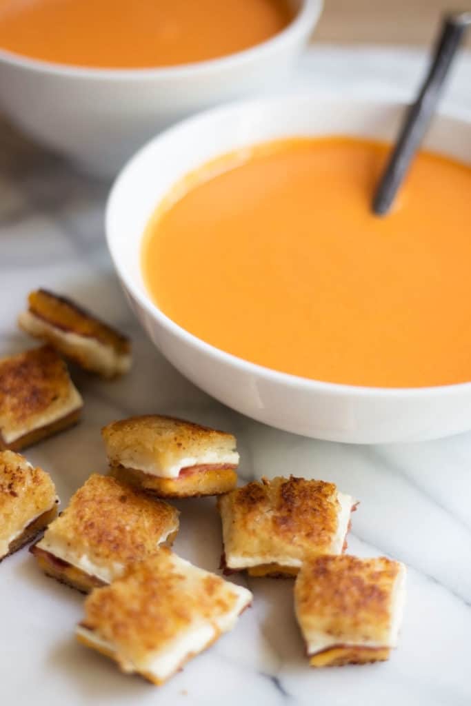 White bowl of tomato soup next to grilled cheese and bacon croutons