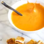 A white bowl of tomato soup with a splash from a crouton being dropped into it surrounded by grilled cheese croutons