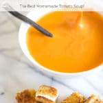 A white bowl of tomato soup with a splash surrounded by grilled cheese and bacon croutons