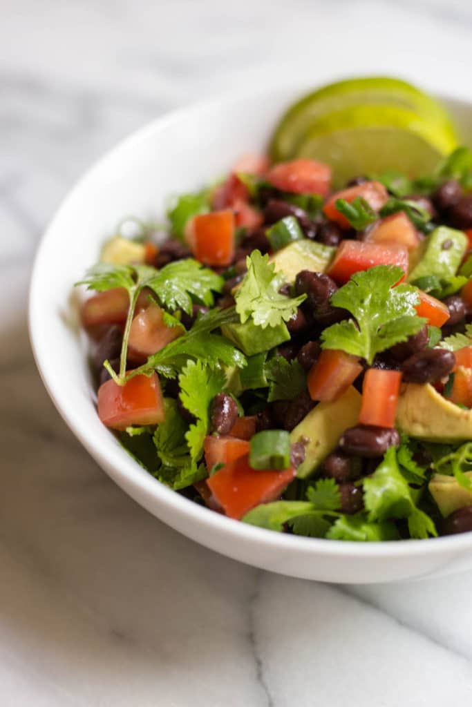 White bowl of salad with chopped avocado, chopped tomatoes, black beans, and cilantro with two slices of lime