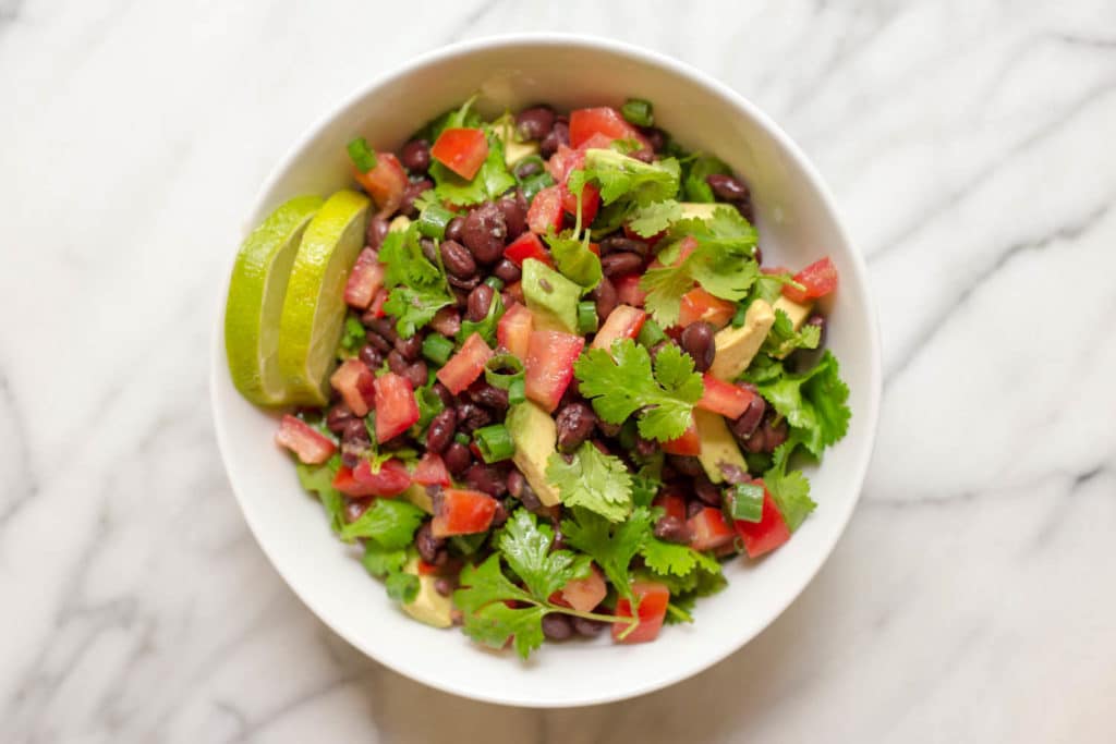 Overhead view of a white bowl of salad with chopped avocado, chopped tomatoes, black beans, and cilantro with two slices of lime