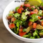 A white bowl of salad with chopped tomatoes, black beans, diced avocado, cilantro and 2 lime slices