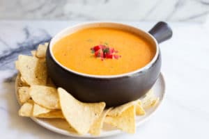 Bowl of spicy queso dip on white plate surrounded with corn chips