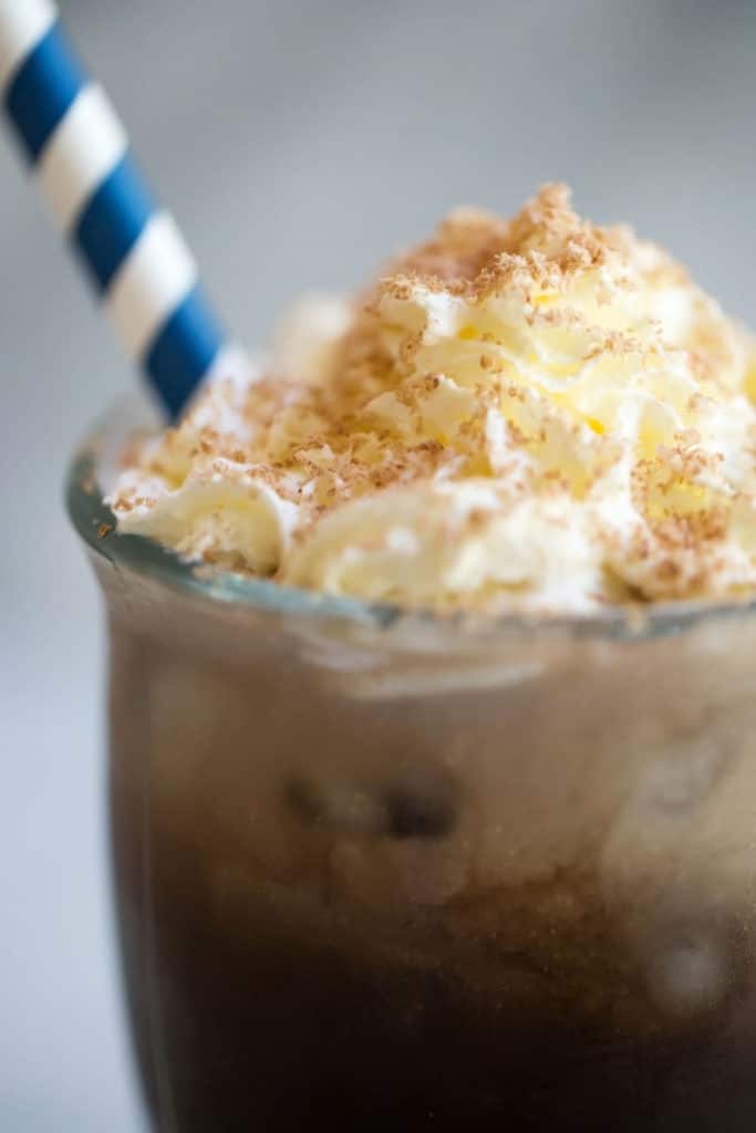 An up close view of a clear mug of iced coffee topped with whipped cream with a blue striped straw
