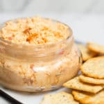 A clear jar filled with pimento cheese surrounded by crackers
