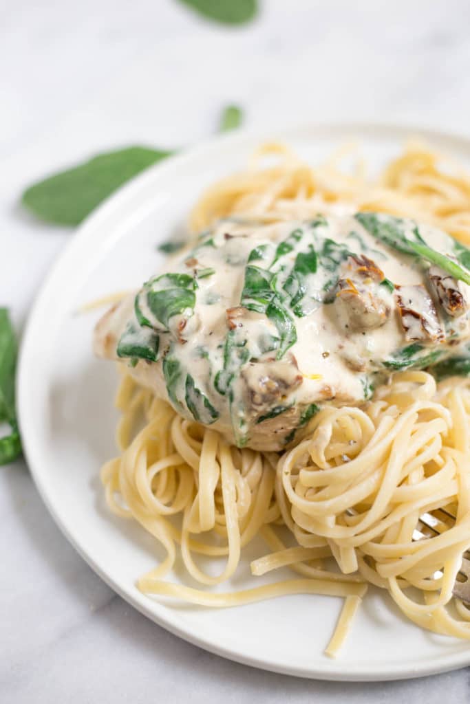 A white plate with linguine pasta topped with a chicken breast covered in white sauce with spinach and sun dried tomatoes