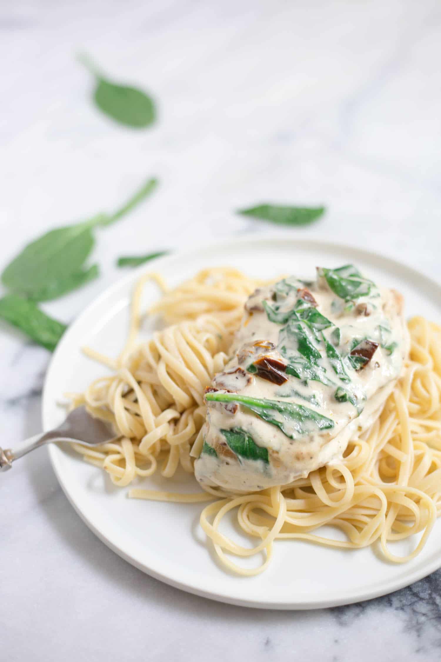 A white plate with linguine pasta and a cooked chicken breast coated with a white sauce with spinach and sun dried tomatoes