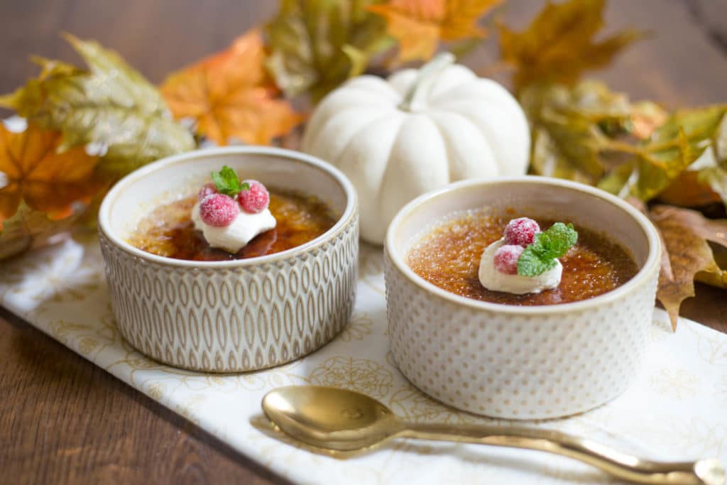 Two ramekins of creme brulee next to a white pumpkin and a gold spoon