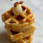 A stack of quartered waffles topped with a scoop of mascarpone, some chopped pecans, drizzled with syrup