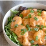 Fried shrimp tossed in a sauce garnished with green onions over a bed of greens in a white bowl