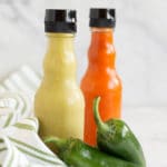 A bottle of green hot sauce next to a bottle of red hot sauce, two jalapenos, and a green striped napkin