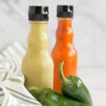 A clear bottle of green hot sauce next to a clear bottle of red hot sauce with 2 jalapeno peppers and a green striped napkin