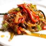 A white plate with julienned red peppers, zucchini, and onions coated in sauce and garnished with white sesame seeds