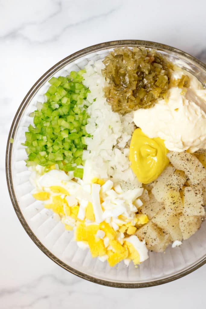 Overhead view of a clear bowl with potatoes, chopped hard boiled eggs, diced onions, chopped celery, relish, mayonnaise and mustard