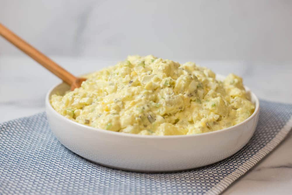 A white bowl of potato salad with a wooden spoon on a blue napkin