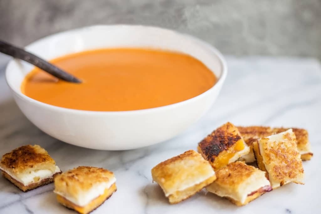 White bowl of tomato soup surrounded by grilled bacon and cheese croutons