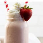 A mason jar filled with a strawberry milkshake topped with whipped cream, a strawberry on the side of the jar, and 2 red striped straws