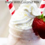 A mason jar filled with strawberry milkshake topped with whipped cream, a strawberry on the side, and two red striped straws