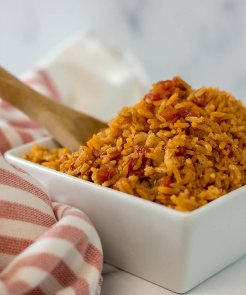 A wooden spoon in a white bowl filled with Mexican rice beside a red striped napkin