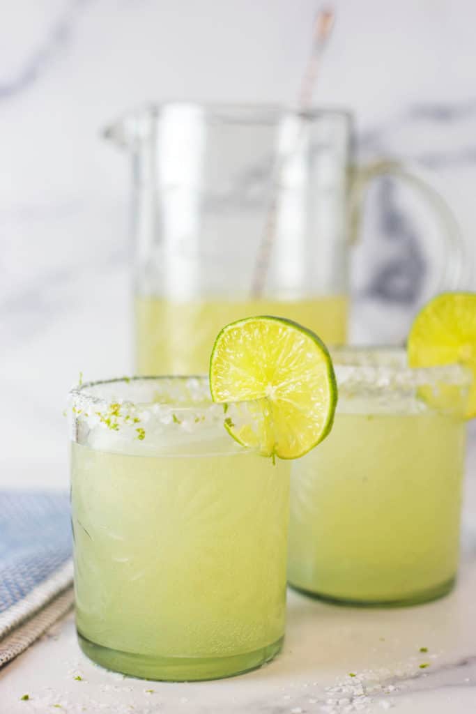 Two glasses of margaritas with slices of lime on the rim with a pitcher of margaritas in the background