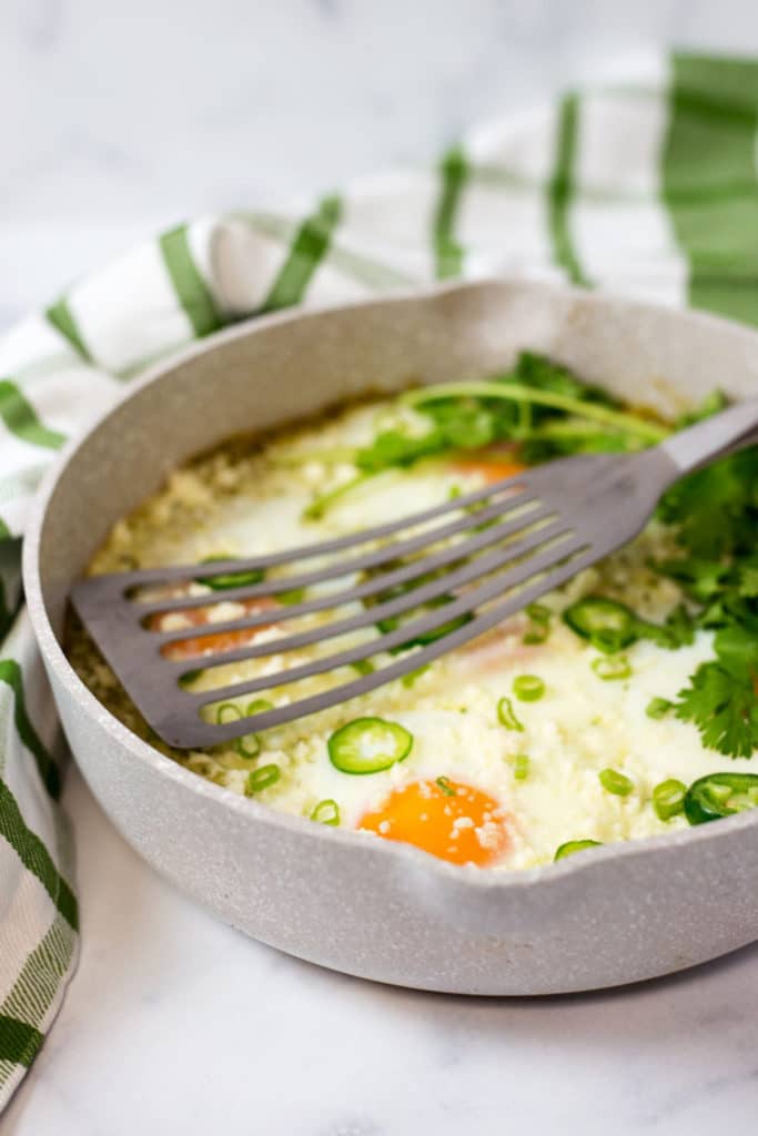 A spatula in a pan of baked eggs garnished with sliced jalapenos and cilantro