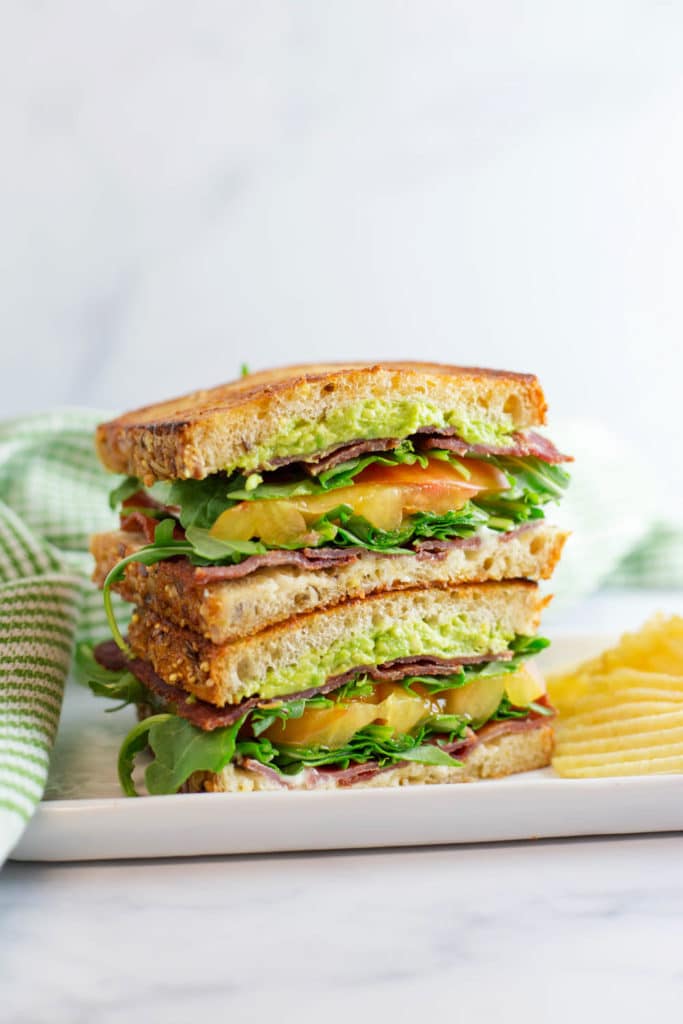 BLT cut in half and stacked on a white plate with a green striped napkin next to potato chips