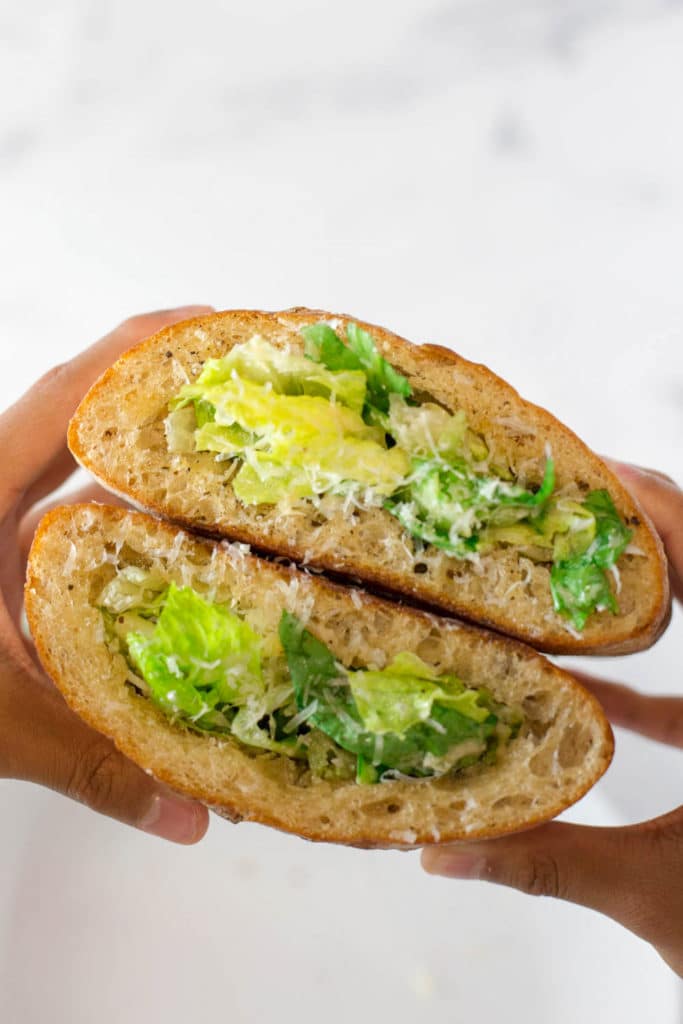 Fingers holding two halves of a ciabatta loaf cut in half, hollowed out, and filled with caesar salad