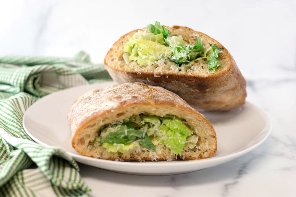 A ciabatta roll, hollowed and filled with caesar salad on a white plate next to a green striped napkin