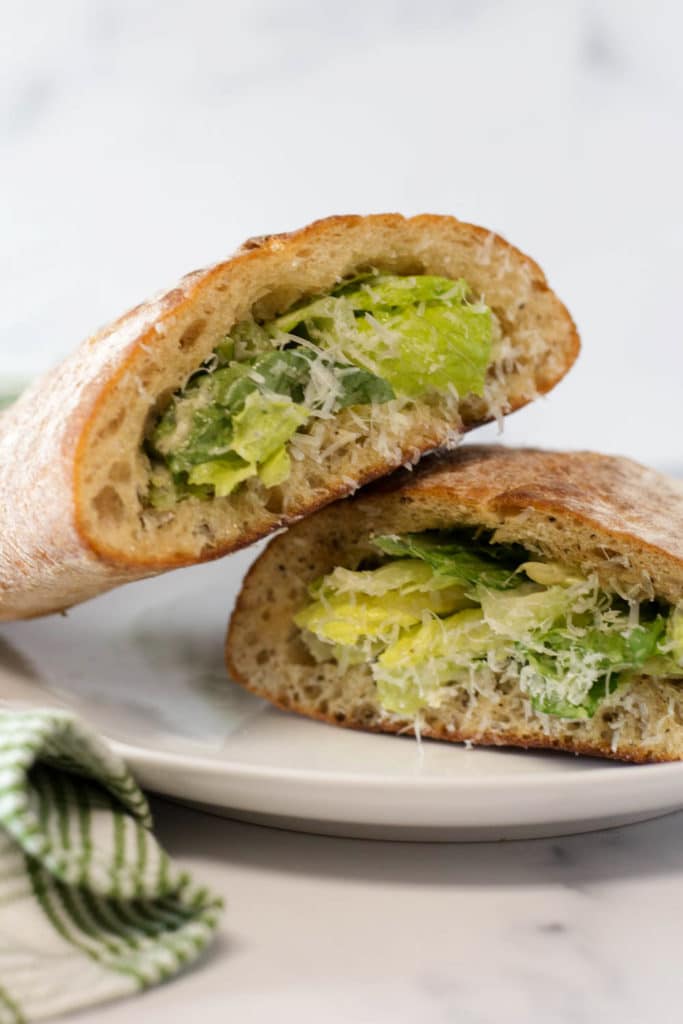 Two halves of a caesar salad sandwich, which is a ciabatta loaf stuffed with green salad on a white plate