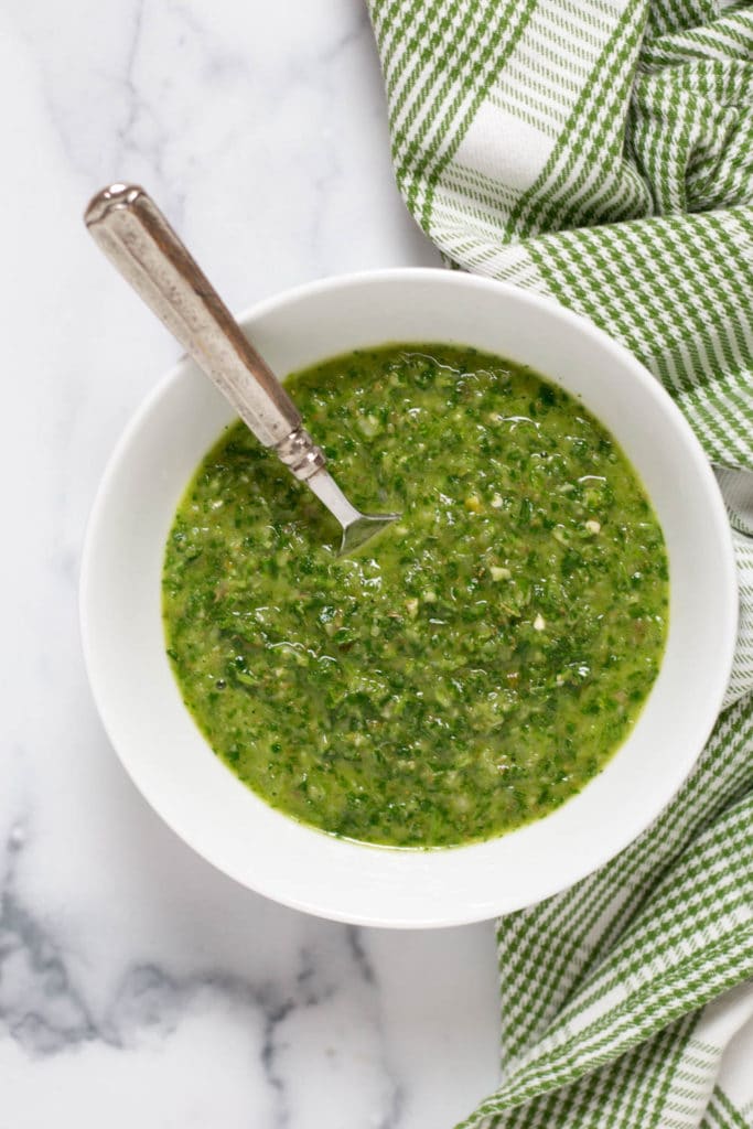 Overhead view of a white bowl of green chimichurri sauce with a silver spoon next to a green checked napkin
