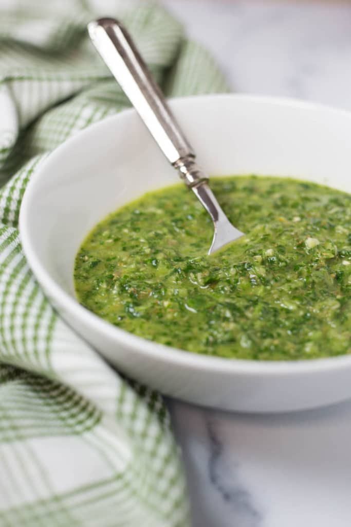 A white bowl of green chimichurri sauce with a silver spoon next to a green checked napkin