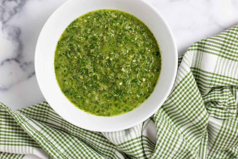 Overhead view of a white bowl of green chimichurri sauce next to a green checked napkin