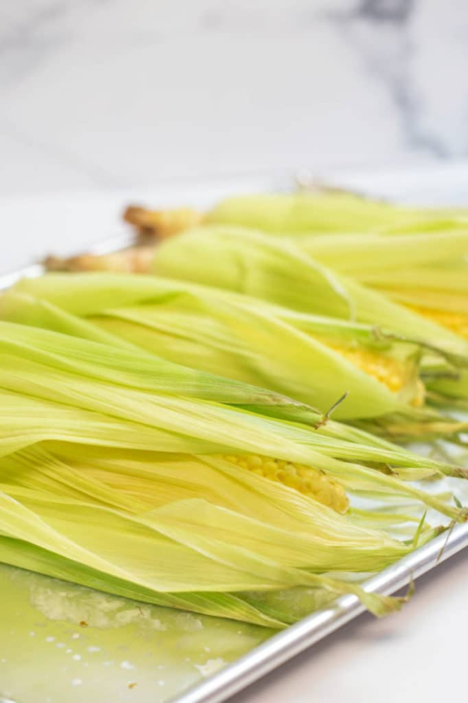 4 ears of corn covered with husks on a baking sheet