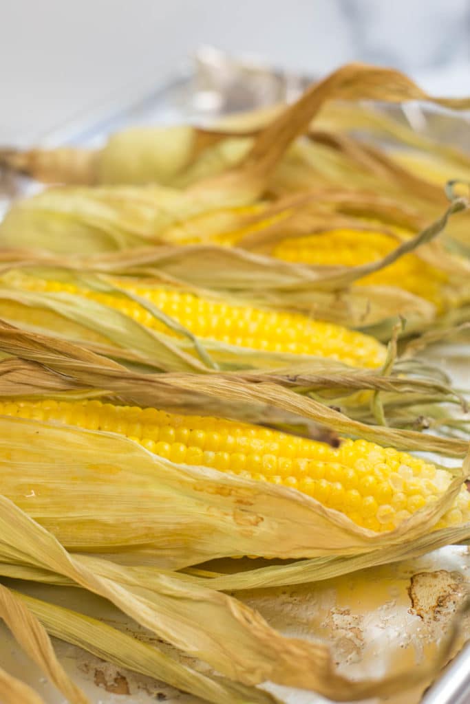 4 ears of corn covered with baked husks on a baking sheet