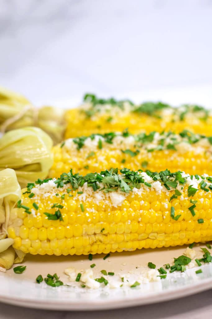 Up close view of an ear of corn on the cob garnished with cotija cheese, cilantro, and chives on a white background