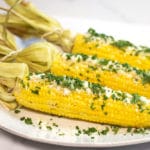 A white plate with 3 ears of corn on the cob garnished with cilantro and cotija cheese