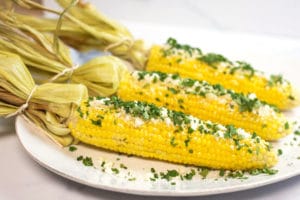 A white plate with 3 ears of corn on the cob garnished with cilantro and cotija cheese