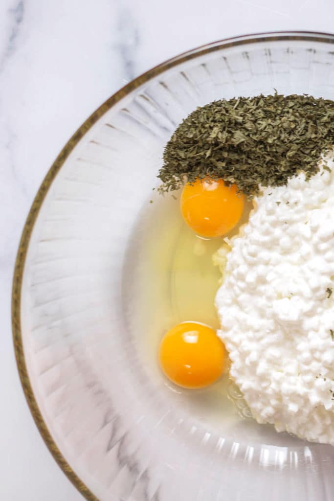 Overhead view of a clear bowl with 2 raw eggs, cottage cheese, and dried parsley