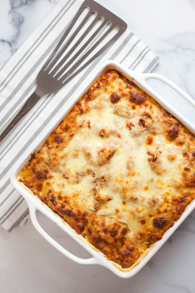 Overhead view of a white baking dish of baked lasagna next to a gray striped napkin topped with a silver spatula