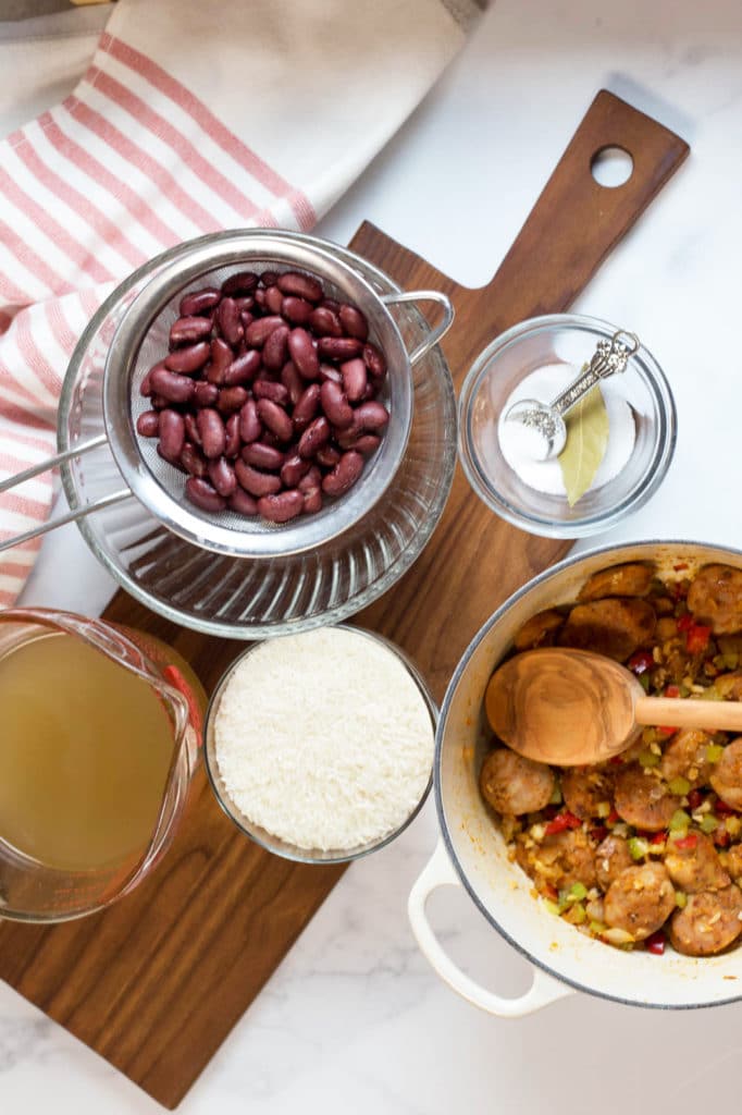 Overhead view of white pot with sausage, onion, celery, bell pepper, and a wooden spoon next to bowls of salt, kidney beans, rice, and broth