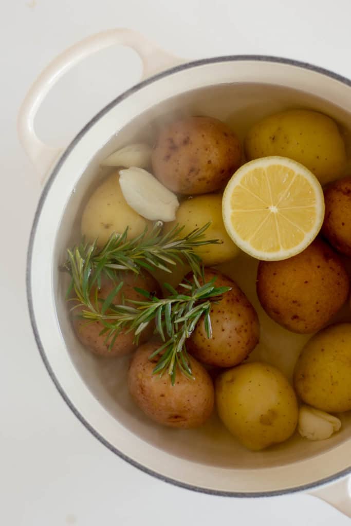 Overhead view of a white pot filled with baby yukon gold potatoes, half a lemon, 2 sprigs of rosemary, and 3 cloves of smashed garlic