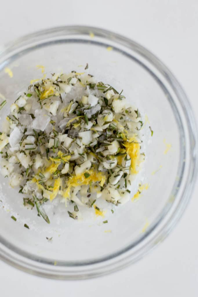 Overhead view of a clear bowl with minced garlic, rosemary, lemon zest, and salt