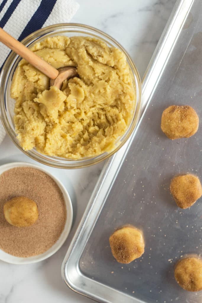Overhead view of a bowl of cookie dough, a ball of unbaked cookie dough in a plate of cinnamon sugar, and a baking sheet with balls of cookie dough