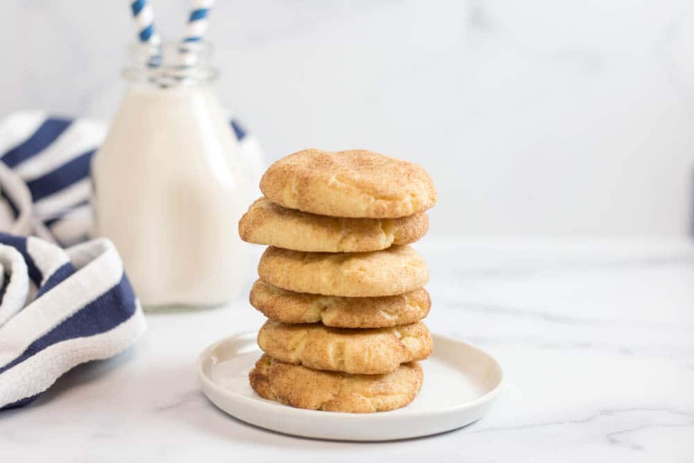 A stack of 6 snickerdoodle cookies on a small round white plate with a clear bottle of milk with blue striped straws and a blue striped napkin in the background