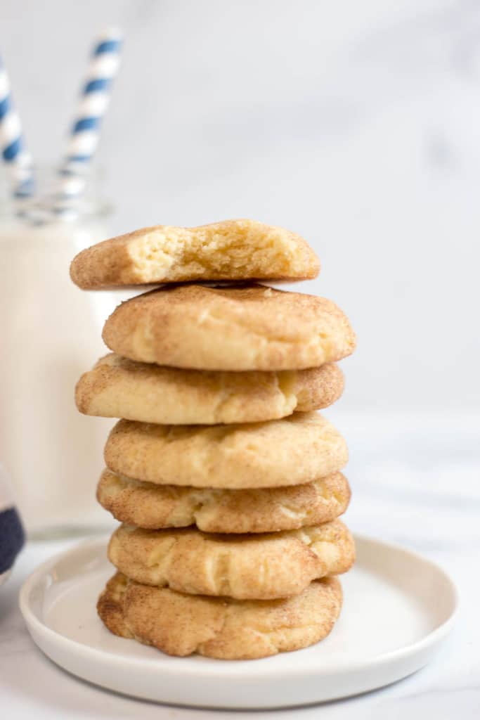 A stack of 6 snickerdoodle cookies on a small round white plate with a clear bottle of milk with blue striped straws in the background